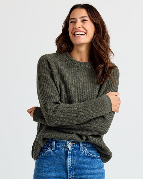 Three-quarter view image of a woman wearing dark olive ribbed cashmere sweater and blue jeans, smiling with arms crossed