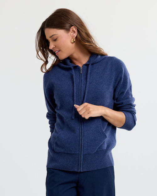Woman wearing indigo zip-up cashmere hoodie with pockets and drawstrings, looking down and smiling