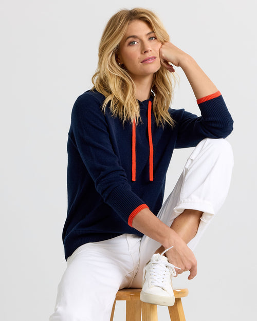 LIFESTYLE / EDITORIAL: Woman seated on stool wearing navy cashmere hoodie with orange accents and white pants