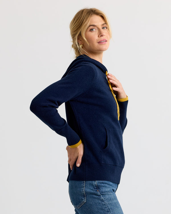 LIFESTYLE / EDITORIAL: Woman in navy and yellow cashmere hoodie, side view, wearing blue jeans against plain background