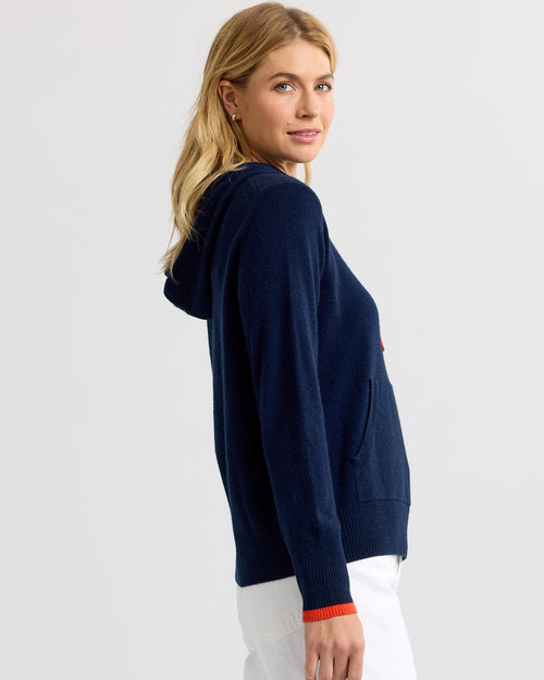 Woman in navy cashmere hoodie with persimmon trim, side view, white pants, plain background