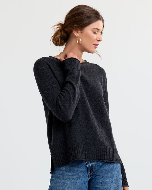 Three-quarter view image of a woman wearing a charcoal cashmere sweater with blue jeans, hand near neck, against plain