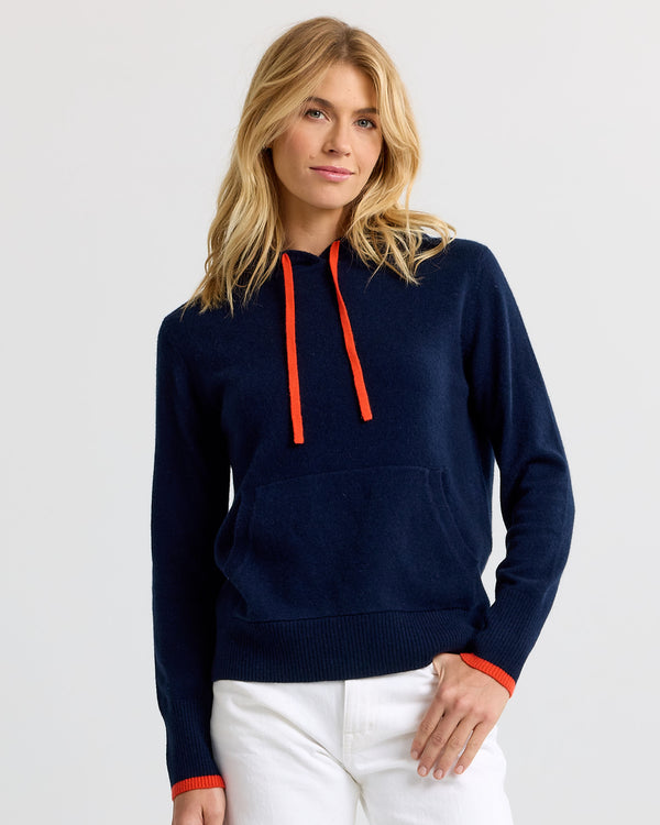 LIFESTYLE / EDITORIAL: Woman wearing navy cashmere hoodie with orange drawstrings and white pants against plain background