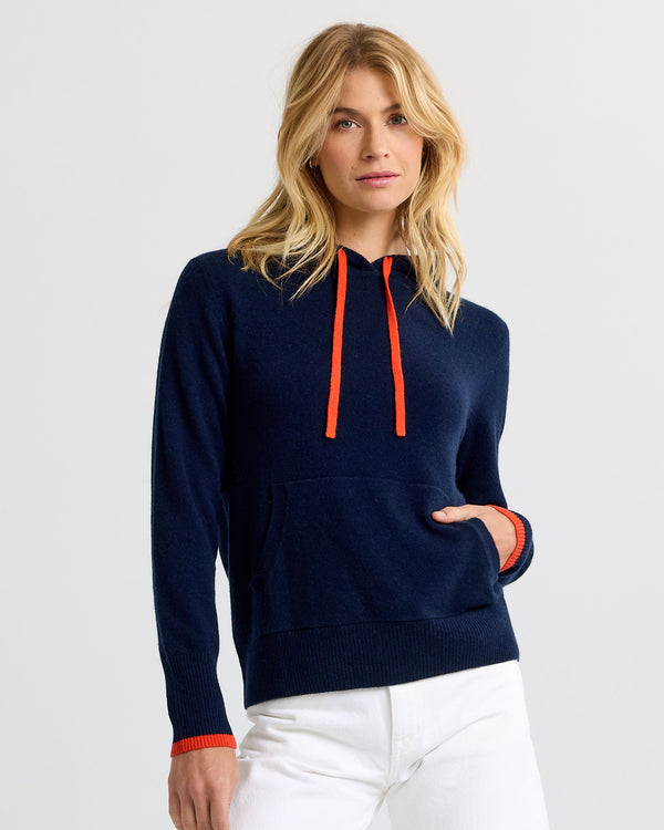 Woman wearing navy cashmere hoodie with persimmon drawstrings and cuffs, paired with white pants, studio background