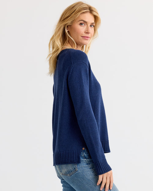 LIFESTYLE / EDITORIAL: Woman wearing navy blue cashmere sweater and jeans, standing sideways against a plain background