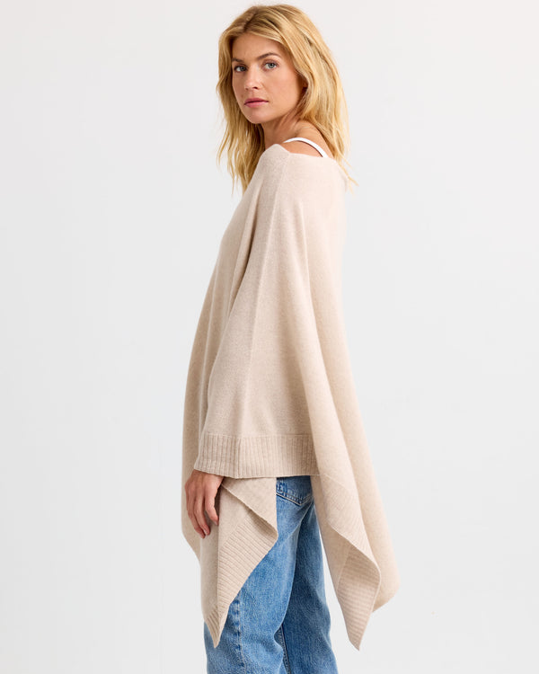 LIFESTYLE / EDITORIAL: Woman wearing oatmeal-colored cashmere poncho and blue jeans, standing sideways against white