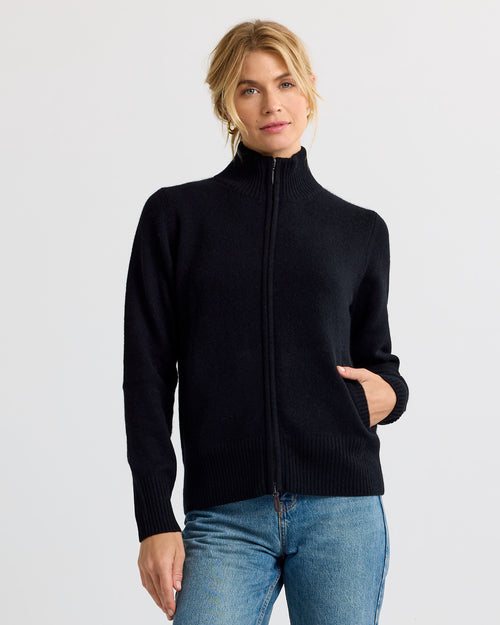 LIFESTYLE / EDITORIAL: Woman wearing black zip-up cashmere sweater and blue jeans, standing against a plain background