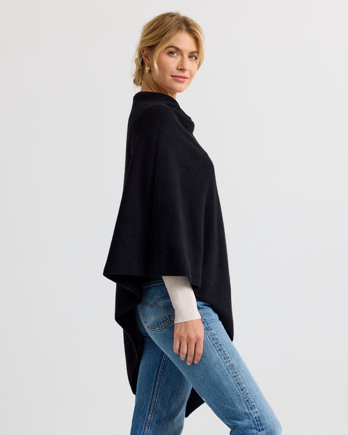LIFESTYLE / EDITORIAL: Woman wearing black cashmere poncho and jeans, standing sideways against a plain background