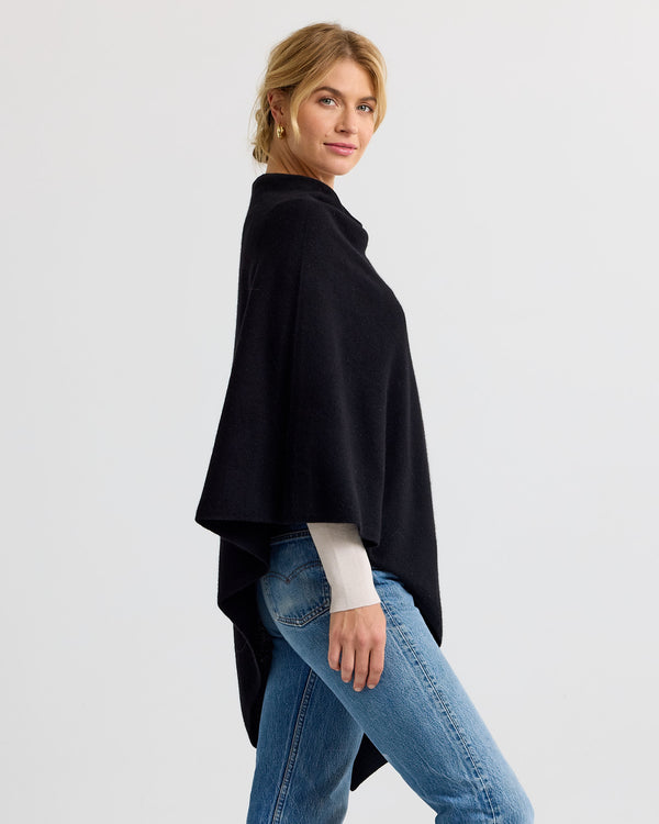 LIFESTYLE / EDITORIAL: Woman wearing black cashmere poncho and jeans, standing sideways against a plain background