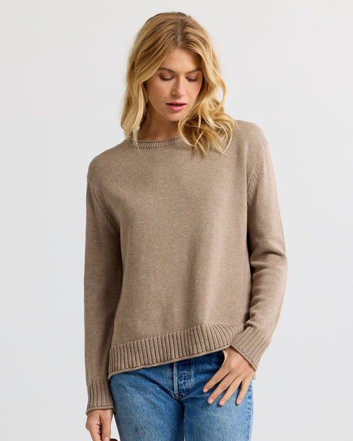 Front view image of a woman wearing a taupe cashmere sweater and blue jeans with her hand in her pocket.