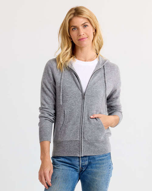 LIFESTYLE / EDITORIAL: Woman wearing grey zip-up cashmere hoodie over white shirt and blue jeans, standing against white