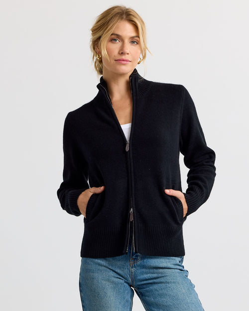 LIFESTYLE / EDITORIAL: Woman wearing black zip-up cashmere sweater with hands in pockets, blue jeans, and gold hoop earrings
