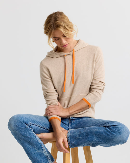 Woman seated cross-legged on stool wearing sand-colored cashmere hoodie with orange drawstrings and blue jeans