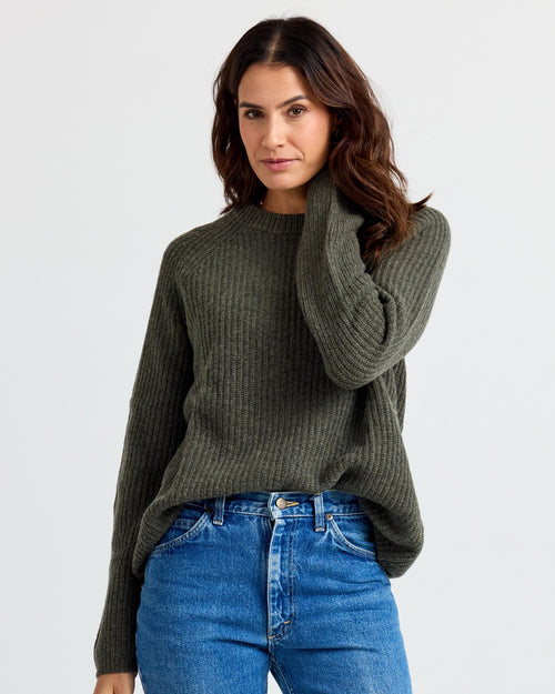 Front three-quarter image of a woman wearing a dark olive ribbed cashmere sweater with blue jeans against white background