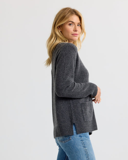 Woman in charcoal cashmere sweater with front pocket, side slits, and blue jeans, facing sideways against plain background