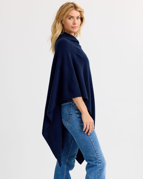 LIFESTYLE / EDITORIAL: Woman wearing navy cashmere poncho and blue jeans, standing sideways against white background
