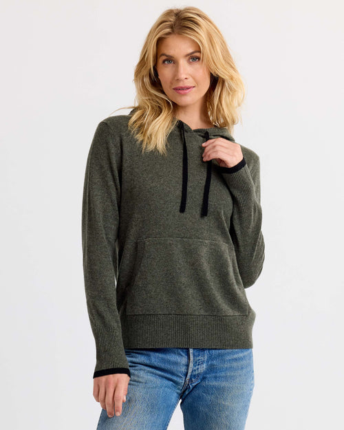 Woman wearing dark olive and black cashmere hoodie with front pocket and blue jeans against white background