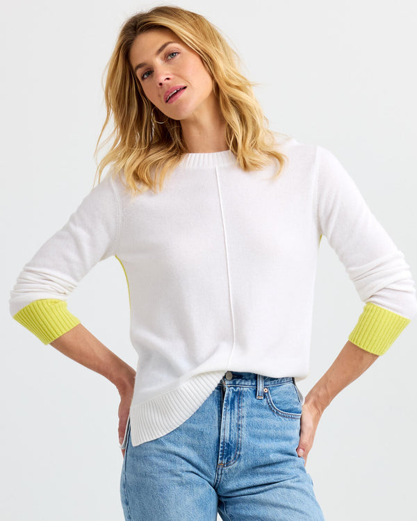 LIFESTYLE / EDITORIAL: Woman wearing white cashmere sweater with yellow cuffs and blue jeans against plain background