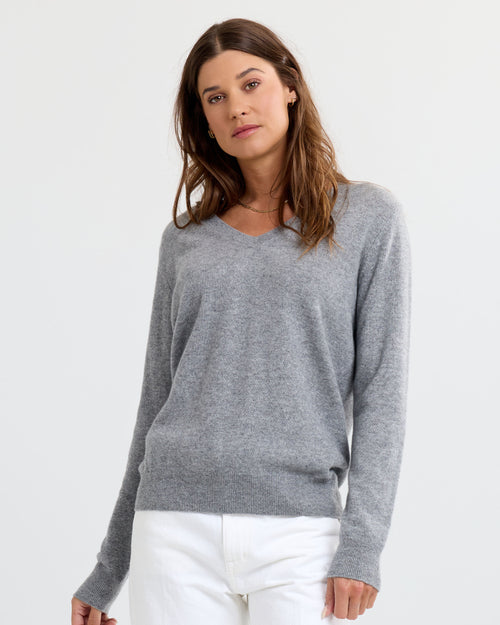 Front-facing image of a woman wearing a grey V-neck cashmere sweater and white pants against plain background