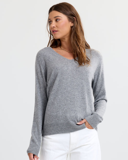 Three-quarter view image of a woman wearing grey V-neck cashmere sweater and white pants, looking to the side