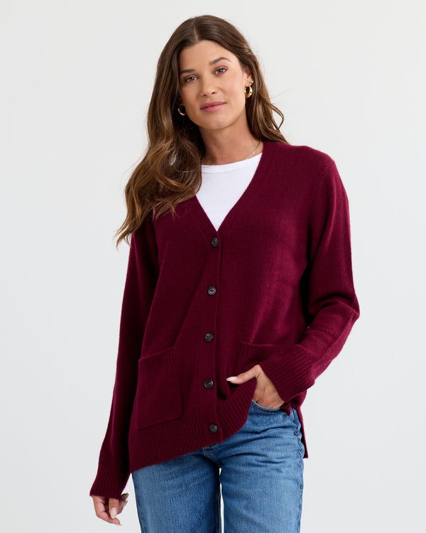 Front view image of a woman wearing a cabernet cashmere cardigan, white top, and blue jeans with hand in pocket.