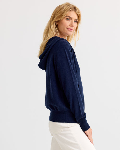 LIFESTYLE / EDITORIAL: Woman wearing navy cashmere hoodie and white pants, standing sideways against a plain background