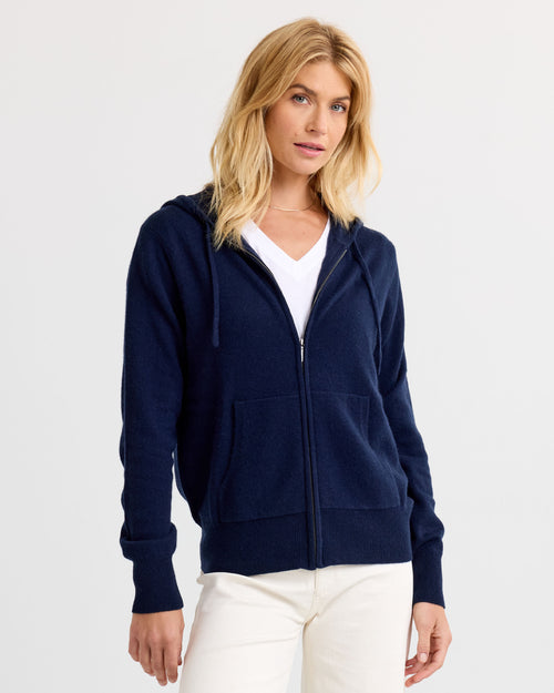 LIFESTYLE / EDITORIAL: Woman wearing navy zip-up cashmere hoodie and white pants against plain background