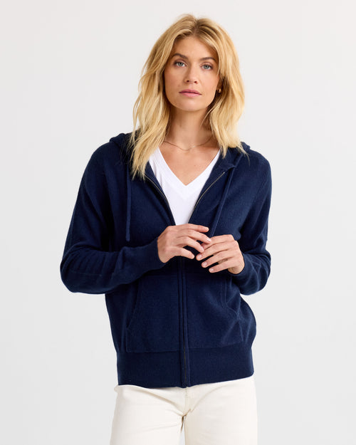 LIFESTYLE / EDITORIAL: Woman wearing navy zip-up cashmere hoodie over white V-neck shirt, standing against white background