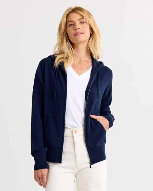 LIFESTYLE / EDITORIAL: Woman wearing navy zip-up cashmere hoodie over white V-neck shirt and cream pants against white