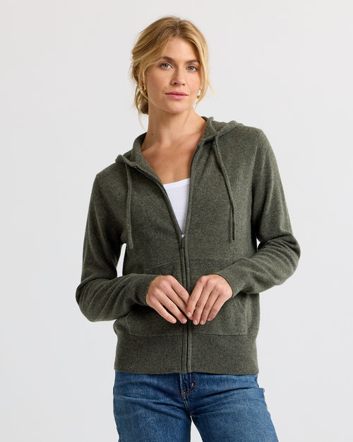 Woman wearing dark olive zip-up cashmere hoodie over white top and blue jeans against plain background
