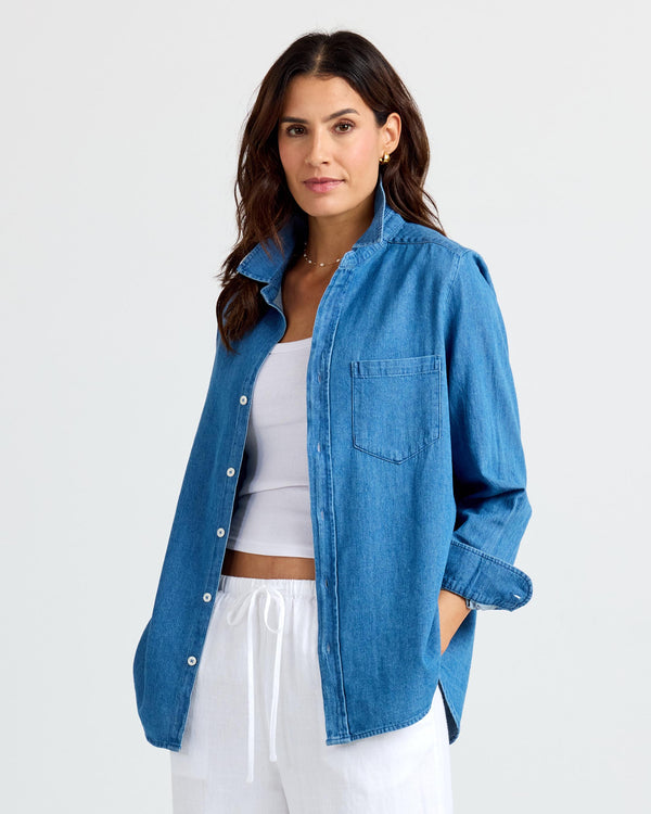 Woman wearing medium wash denim shirt over white tank and white drawstring pants against plain background