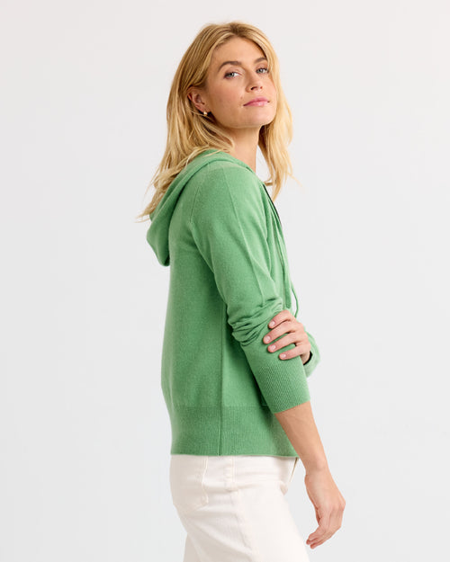 LIFESTYLE / EDITORIAL: Woman in green cashmere hoodie and white pants standing sideways against plain background