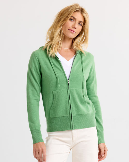 Woman wearing green zip-up cashmere hoodie over white top and cream pants against plain background