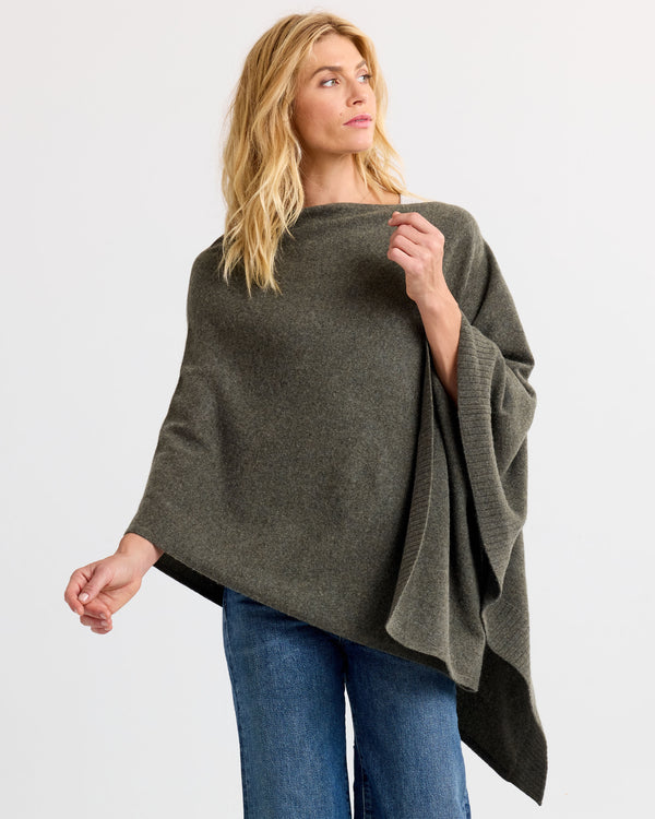 LIFESTYLE / EDITORIAL: Woman wearing dark olive cashmere poncho with jeans, standing against a plain white background
