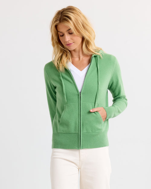 LIFESTYLE / EDITORIAL: Woman wearing a green zip-up cashmere hoodie and white pants against a plain background