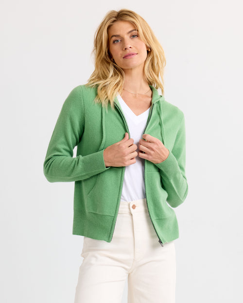 Woman wearing matcha green zip-up cashmere hoodie over white V-neck tee and cream pants against white background