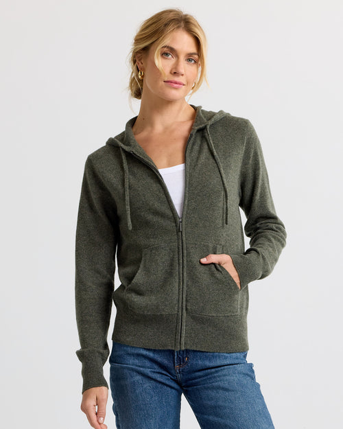 Front view image of a woman wearing a dark olive zip-up cashmere hoodie with pockets and blue jeans against white background