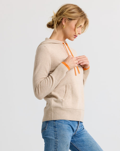 LIFESTYLE / EDITORIAL: Woman in beige cashmere hoodie with orange accents and blue jeans, side profile against white