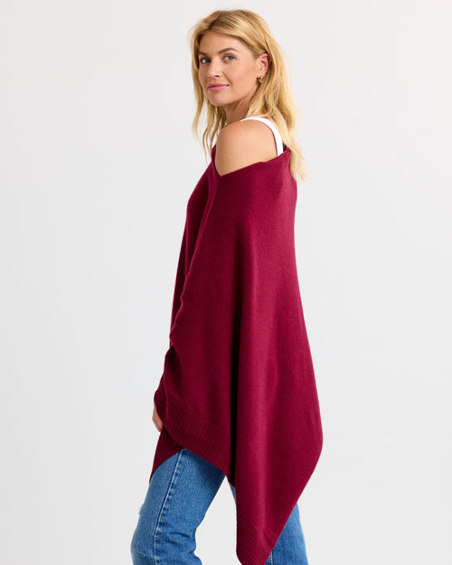 LIFESTYLE / EDITORIAL: Woman wearing off-shoulder barolo red cashmere poncho with jeans, standing against white background