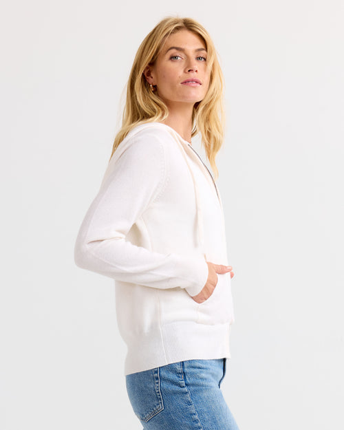 LIFESTYLE / EDITORIAL: Woman wearing ivory cashmere hoodie and blue jeans, standing sideways against white background