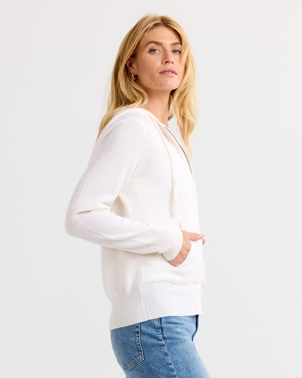 LIFESTYLE / EDITORIAL: Woman wearing ivory cashmere hoodie and blue jeans, standing sideways against white background