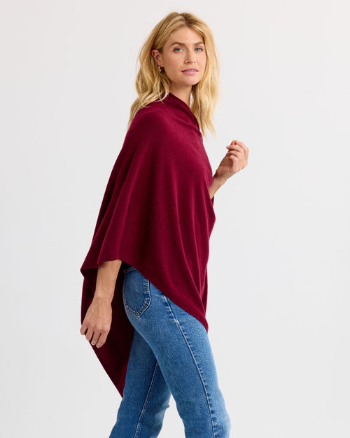 LIFESTYLE / EDITORIAL: Woman wearing a barolo-colored cashmere poncho with blue jeans, standing against a white background