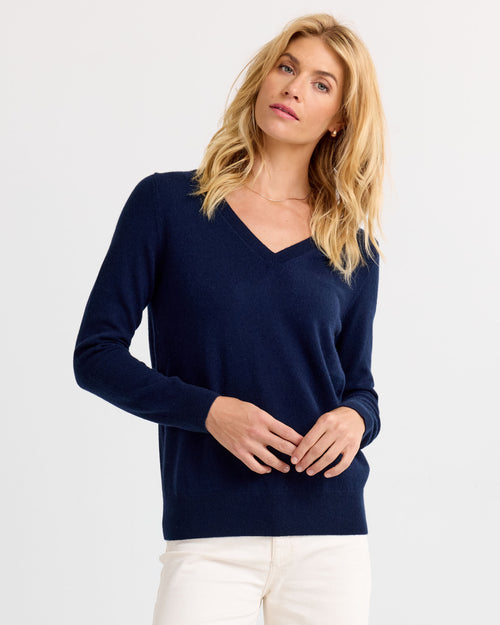 LIFESTYLE / EDITORIAL: Woman wearing navy V-neck cashmere sweater and white pants against a plain background