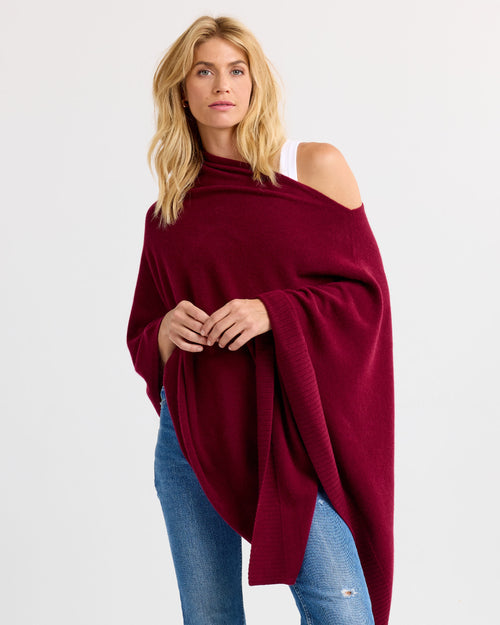 LIFESTYLE / EDITORIAL: Woman wearing a burgundy cashmere poncho over white top and blue jeans against a plain background