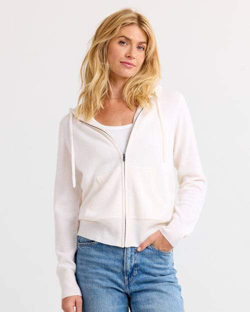 LIFESTYLE / EDITORIAL: Woman wearing ivory zip-up cashmere hoodie and white tank top with blue jeans against white background