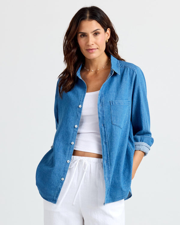 Three-quarter image of a woman wearing a medium wash denim shirt over a white tank and white drawstring pants