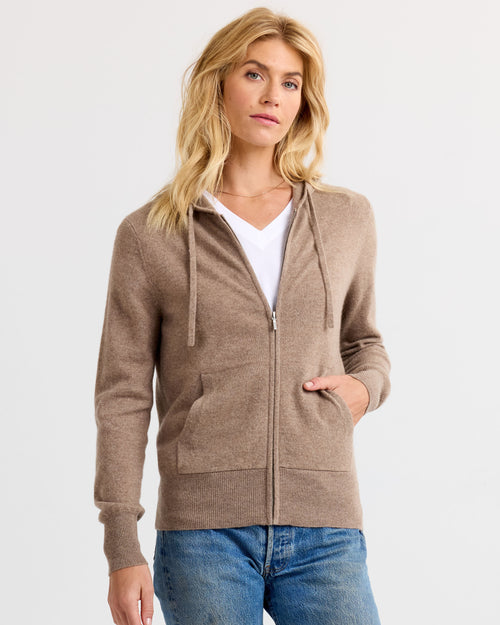 LIFESTYLE / EDITORIAL: Woman wearing driftwood-colored zip-up cashmere hoodie with pockets and blue jeans against white