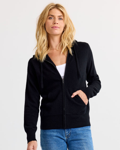 LIFESTYLE / EDITORIAL: Woman wearing black zip-up cashmere hoodie with blue jeans and white top against plain background