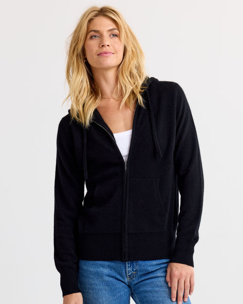 Front-facing image of a woman wearing a black zip-up cashmere hoodie with blue jeans against white background