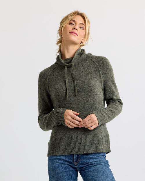 Woman wearing dark olive cashmere turtleneck sweater with drawstrings and blue jeans against white background